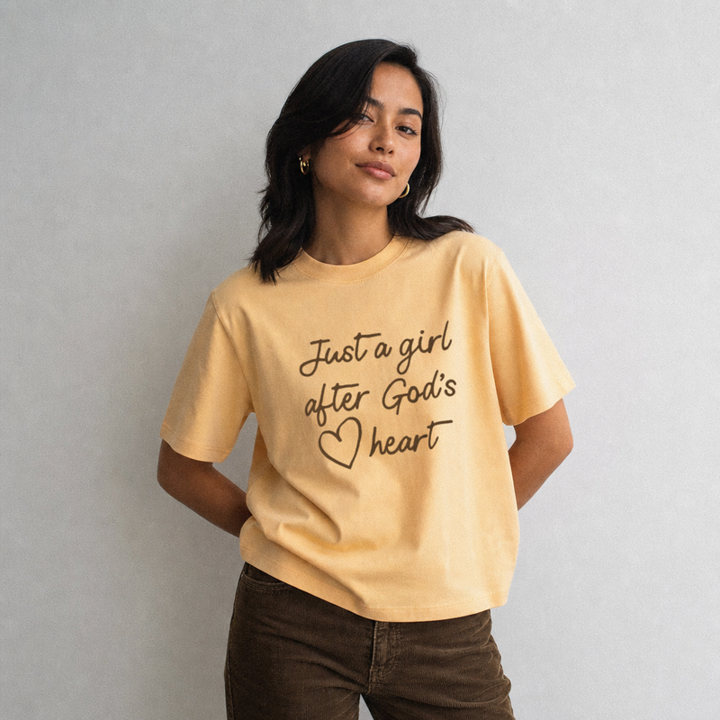A woman wearing a pale yellow graphic t-shirt with the text 'Just a girl after God's heart' in black script, paired with brown pants, standing against a neutral gray wall.