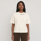 A woman modeling a cream-colored short-sleeve t-shirt with 'After Coco' printed in black script on the chest, paired with brown pants, against a light gray background.