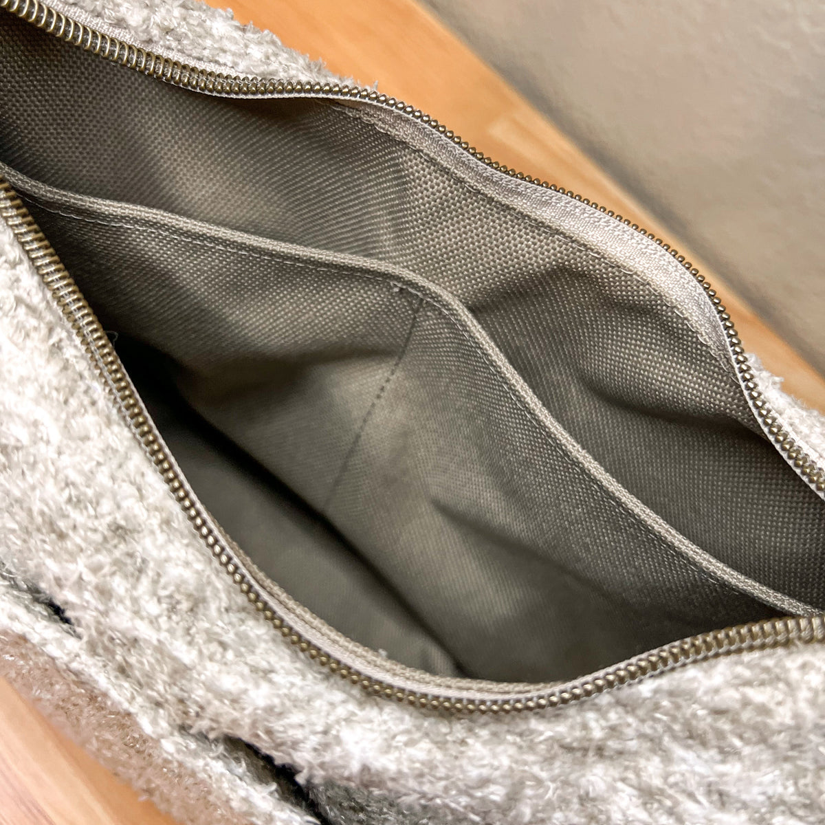 Close-up of the interior of a textured light gray bag, showing a gray fabric lining and a silver zipper, resting on a wooden surface.