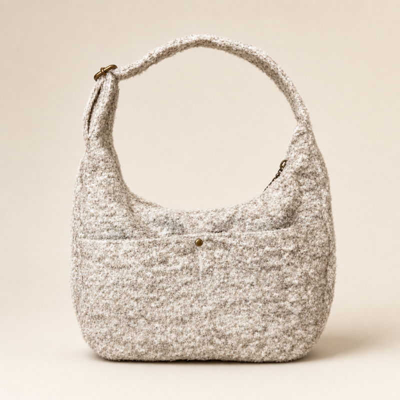 A textured, chunky knit hobo shoulder bag in a speckled beige and white boucle fabric, featuring a single top handle, a front slip pocket with a decorative button, and a zippered main compartment, shown against a solid cream background.