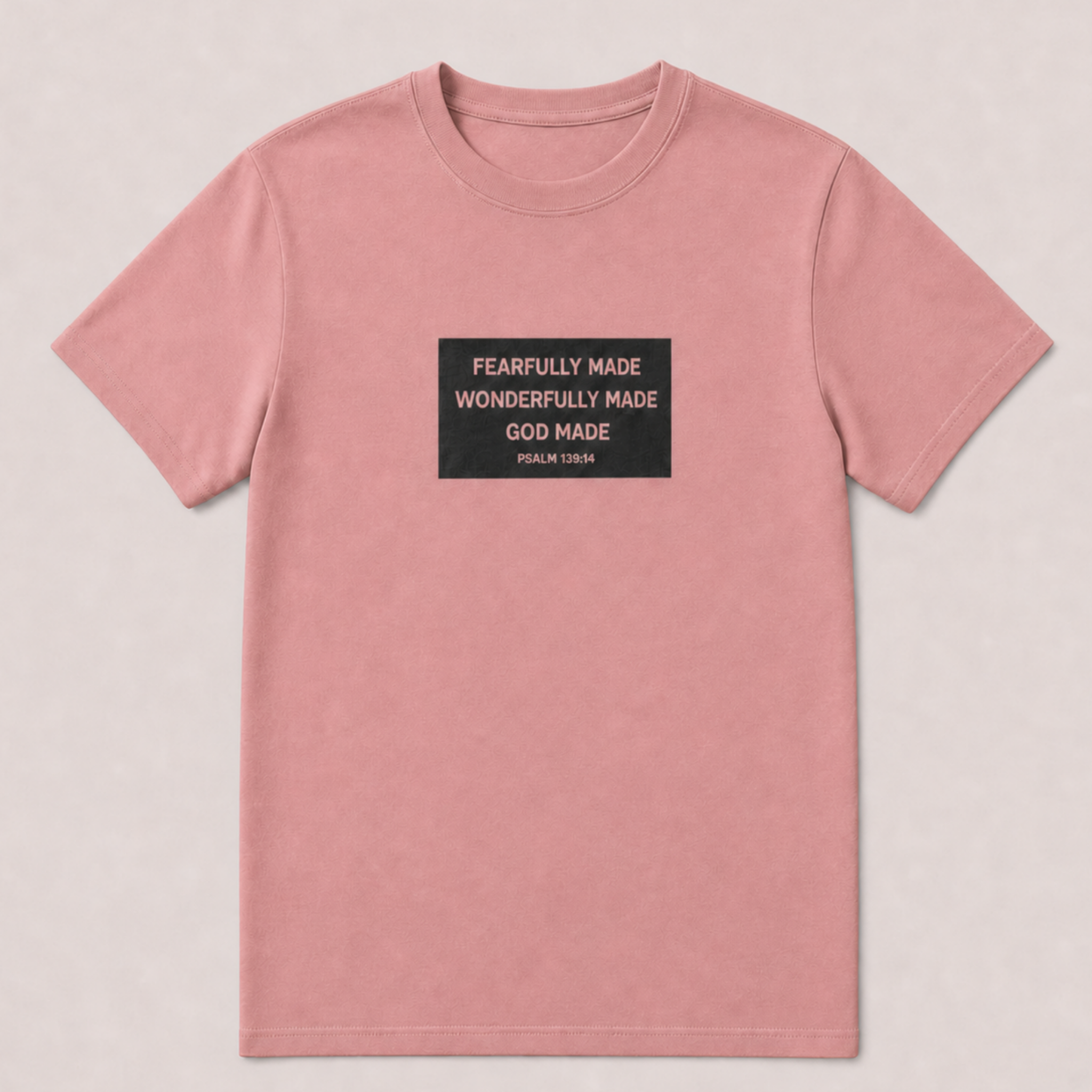 A dusty pink short-sleeve crewneck t-shirt with a black rectangular graphic on the chest containing white text that reads 'FEARFULLY MADE WONDERFULLY MADE GOD MADE PSALM 139:14'. The t-shirt is displayed flat against a light beige textured background.