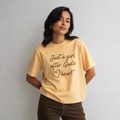 A woman wearing a pale yellow graphic t-shirt with the text 'Just a girl after God's heart' in black script, paired with brown pants, standing against a light gray wall.