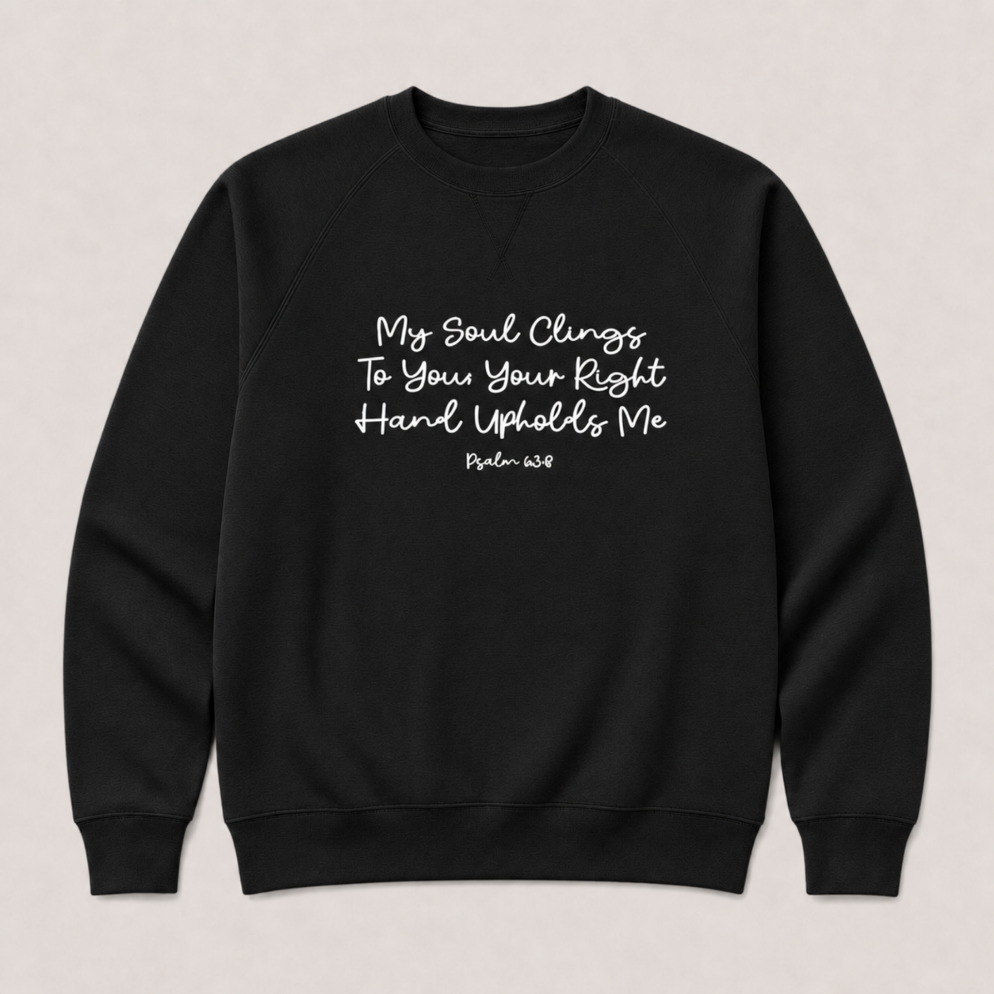 A black crewneck sweatshirt with white printed text on the chest reading 'My Soul Clings To You Your Right Hard Upholds Me Psalm 63:8' on a light beige background.