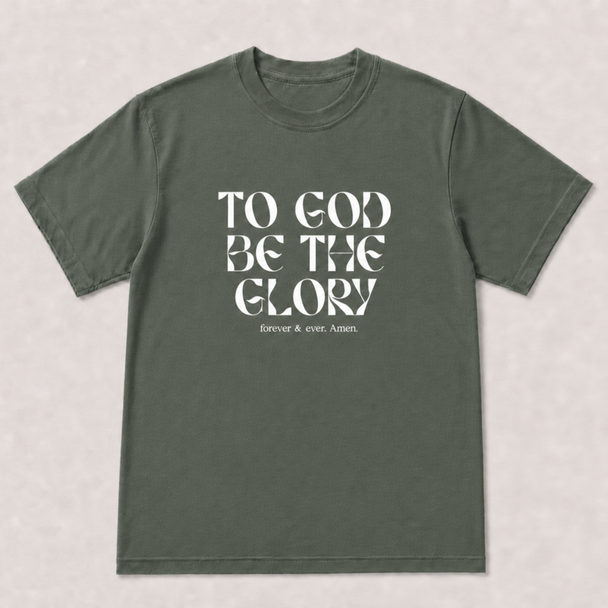 An olive green short-sleeve t-shirt with a white typographic print on the front reading 'TO GOD BE THE GLORY' and 'forever and ever AMEN.' The shirt is laid flat against a light gray textured background.