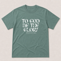 A sage green crewneck t-shirt with the text 'TO GOD BE THE GLORY' printed in white on the front, displayed flat against a light beige background.